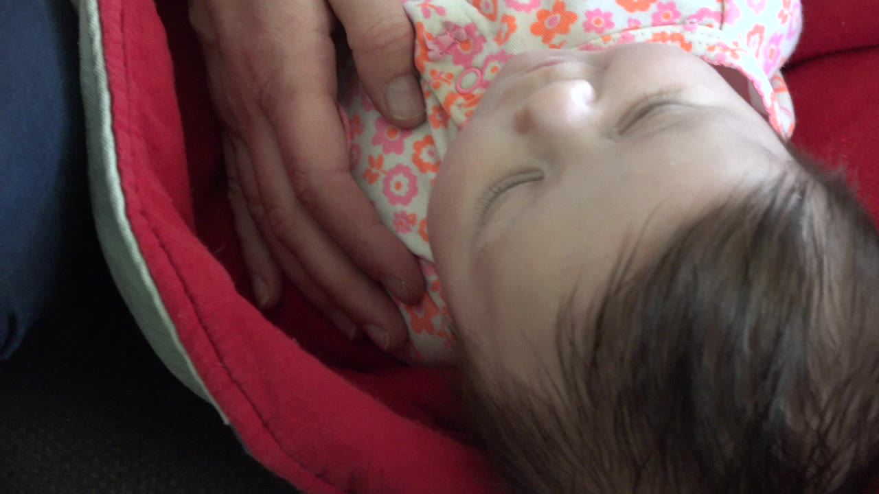 At 0:01 in Westminster, Colorado, the baby girl's face remains central as adult hands cradle her side against the red blanket.