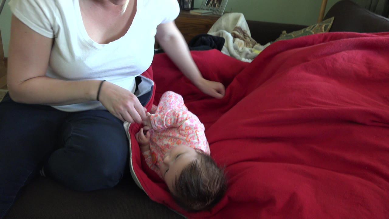 In Westminster, Colorado, a woman in a white shirt sits at the edge of a bed as a baby in a pink sleeper lies on a bright red blanket near her lap.