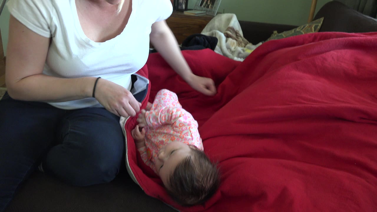 In Westminster, Colorado, the woman leans over the red blanket with one hand near the baby in a pink patterned sleeper while the bedroom background stays visible.
