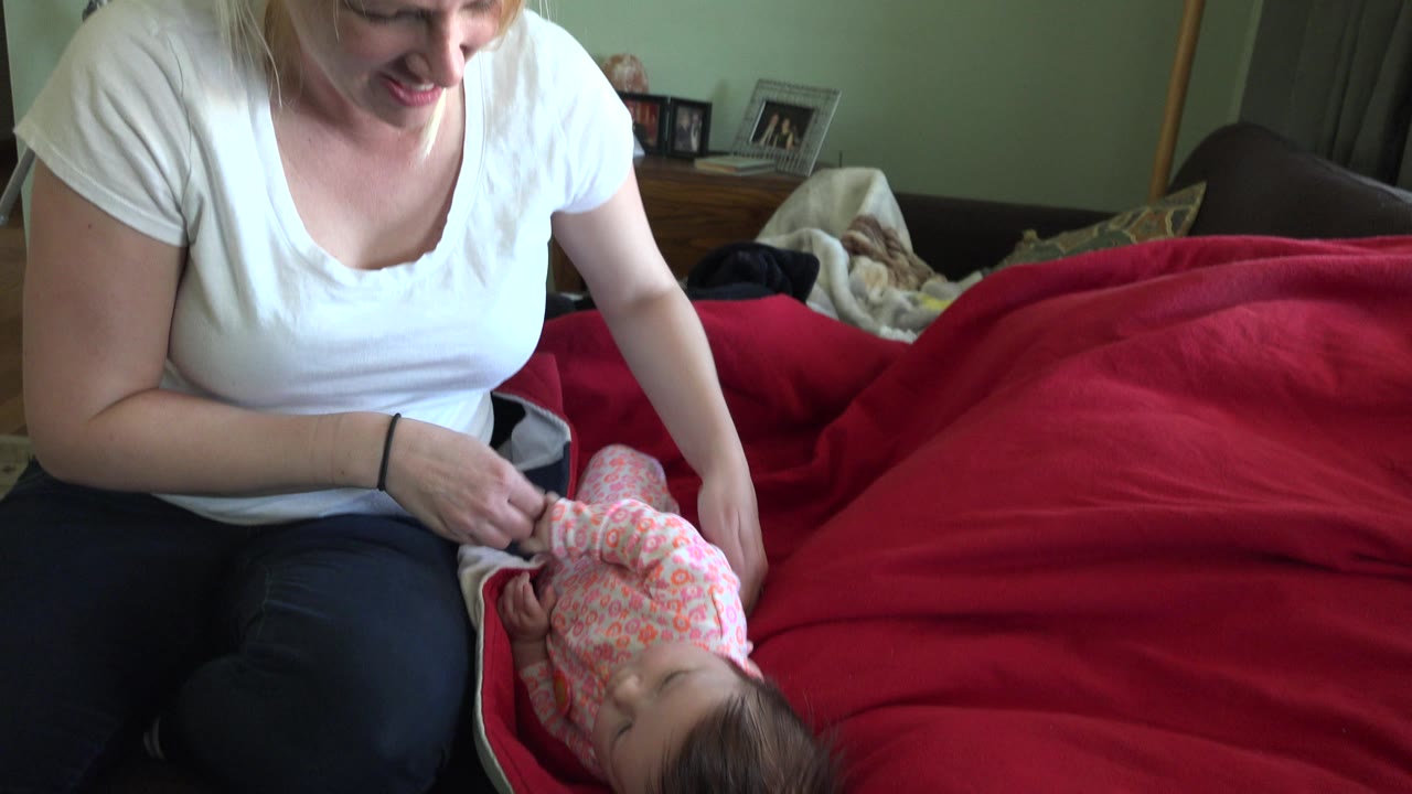 Smiling woman bends toward a baby on a red bed blanket.