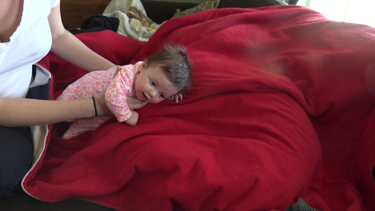 In Westminster, Colorado, the baby turns more sideways across the red bedding as the adult's arm stays along the left edge of the frame.