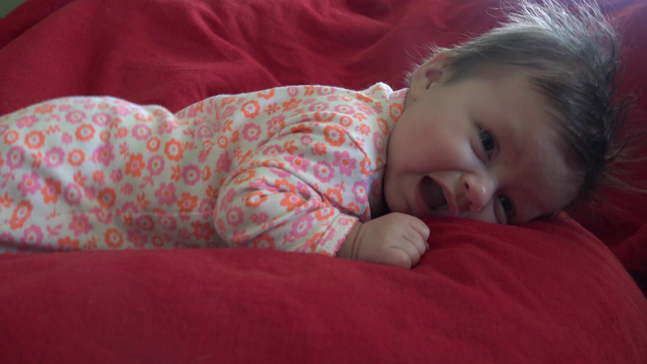 Baby lying face down on red bedding in a floral onesie.