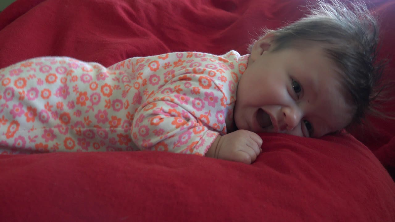 At 0:01, the baby remains prone on red cushions in Westminster, Colorado, with the cheek low to the fabric and the mouth open in a cry.