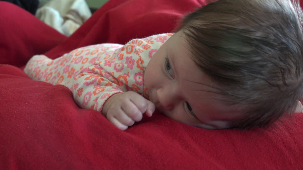Baby lying on red bedding with thumb near mouth.