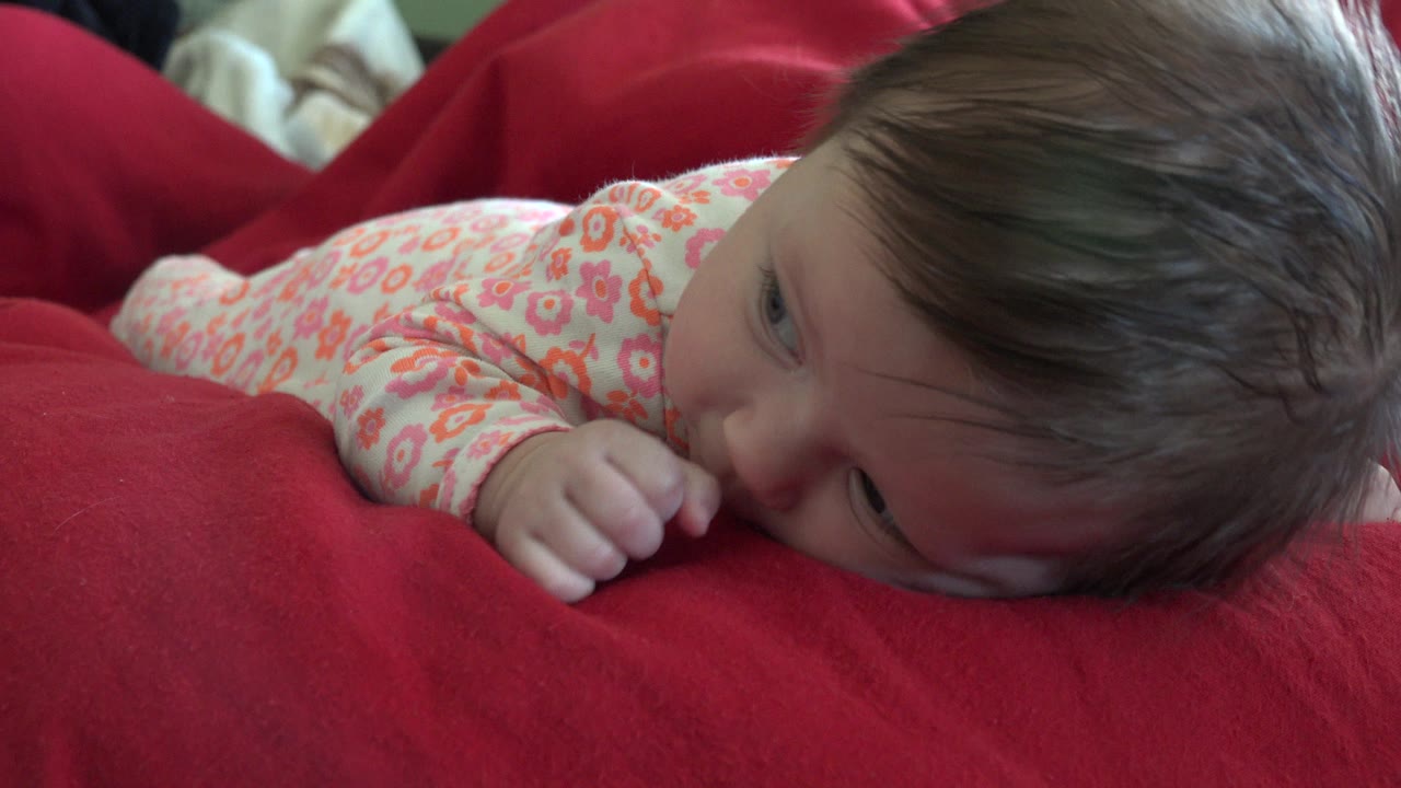 The baby remains face-down on the red bed in Westminster, Colorado, with one eye partly open and a hand tucked near the mouth.