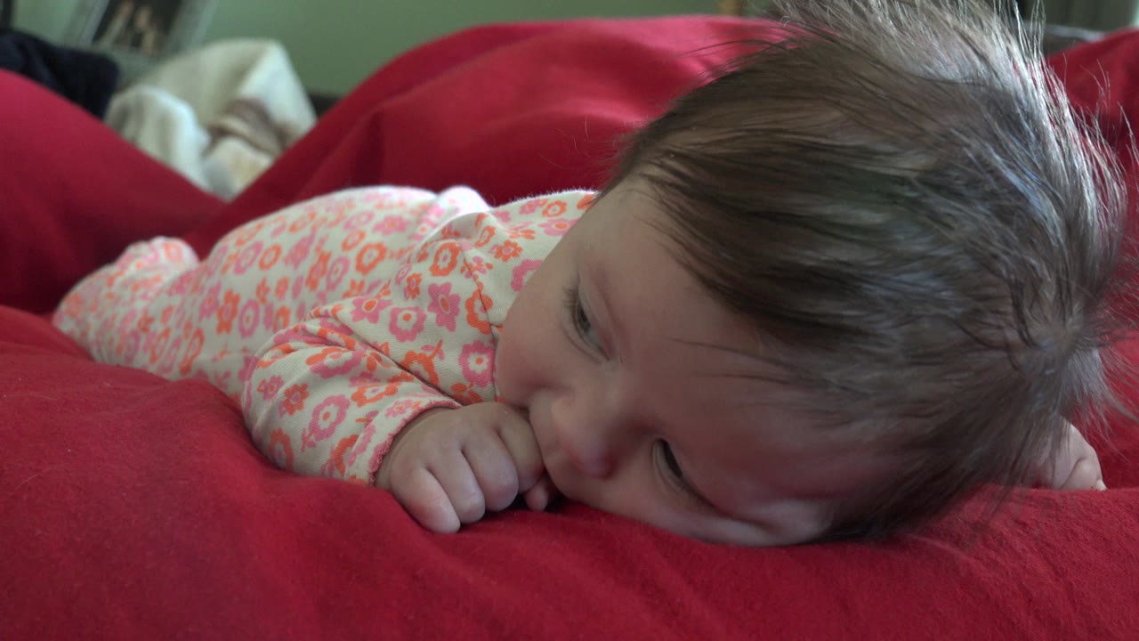 Infant lying on red bedding with thumb near mouth.