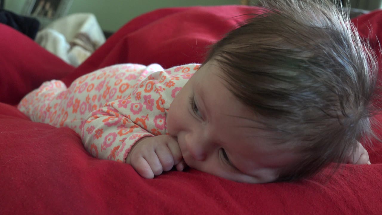 At 0:01, the infant remains on her stomach in a close side view, wearing a pale floral outfit while her cheek and hand rest near the red blanket.