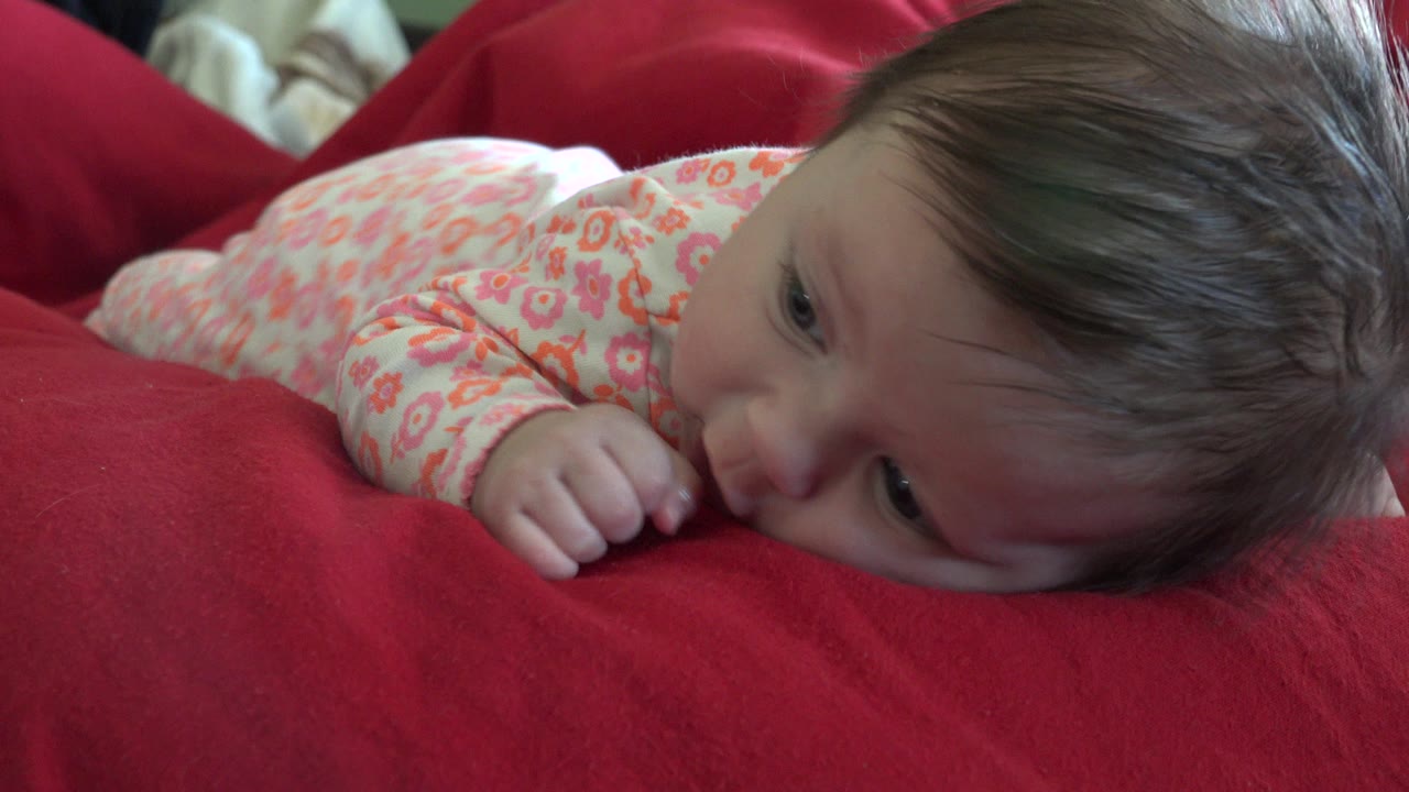 The baby remains low on the red blanket in Westminster, Colorado, with the face turned sideways and the floral sleeve visible near the foreground.