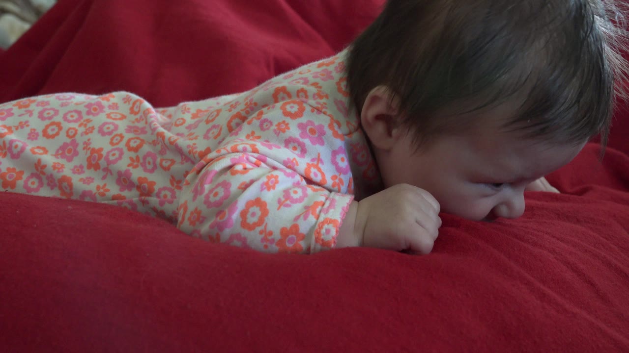 In Westminster, Colorado, a baby in a pink floral outfit lies face-down with one hand tucked near her mouth on a red blanket.