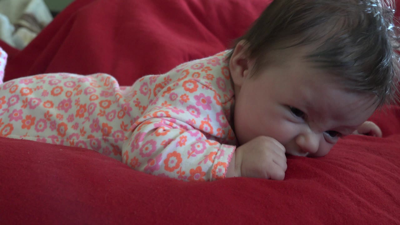 The infant remains low against the red blanket in Westminster, Colorado, with the floral sleeve and cheek visible in soft indoor light.