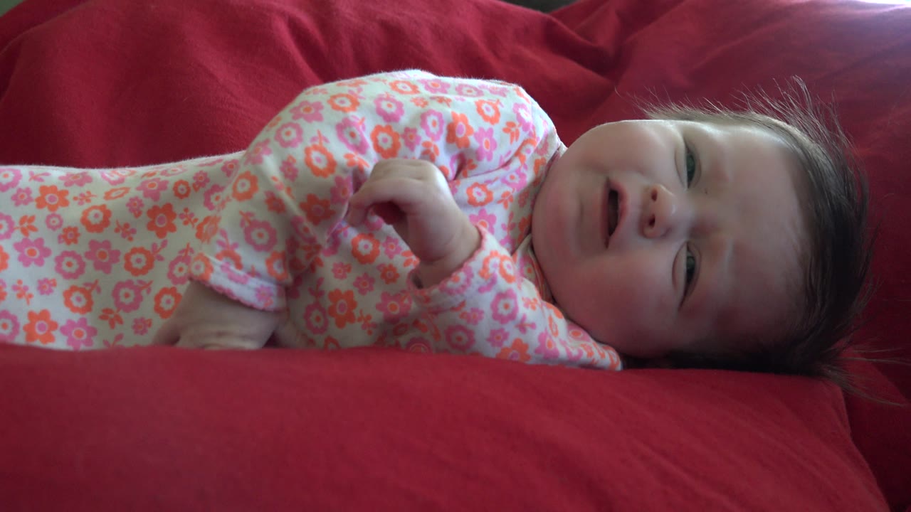 A young child in a floral outfit lies across the red couch in Westminster, Colorado, with one fist lifted near the chest and face turned toward camera.