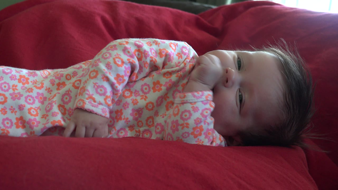 At the start of the Westminster, Colorado clip, a baby in a pink floral onesie lies on a red couch with one hand near the mouth and the face turned toward the camera.