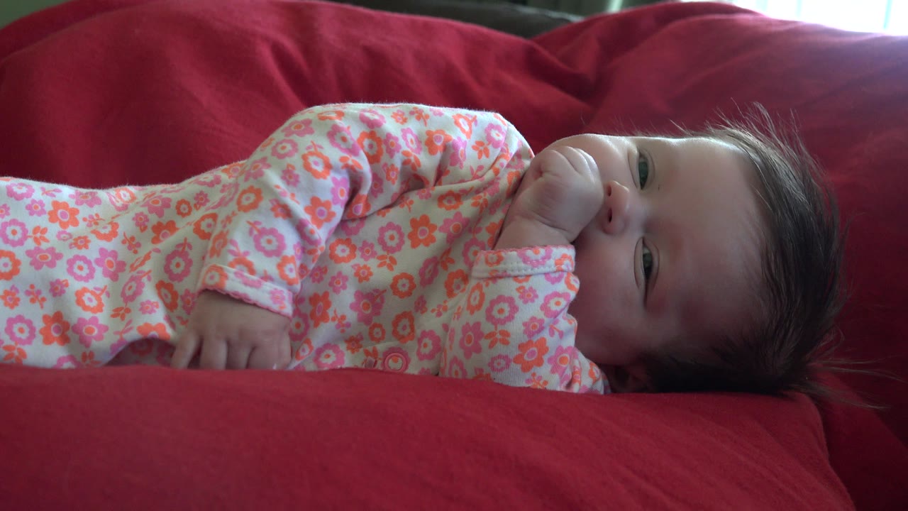 One second in, the baby remains sideways on the red couch in Westminster, Colorado, with the face visible and the fist held close to the mouth.