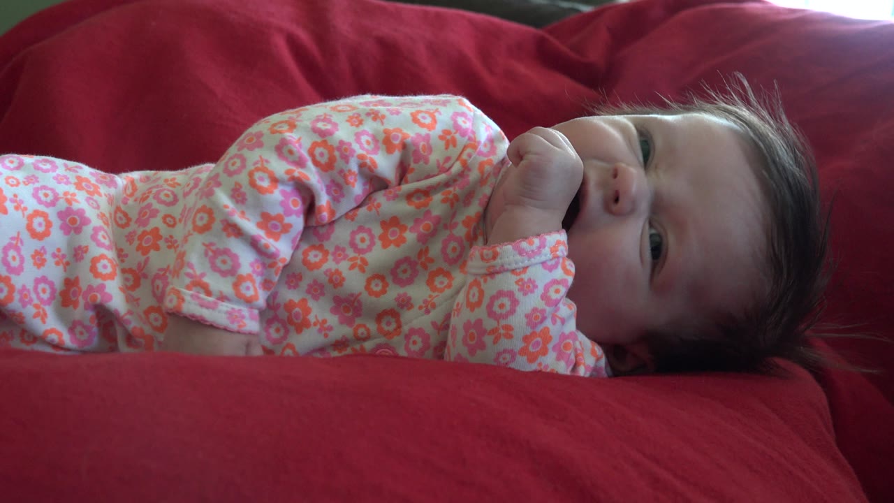Baby lying on a red couch with arm bent and face partly turned upward.