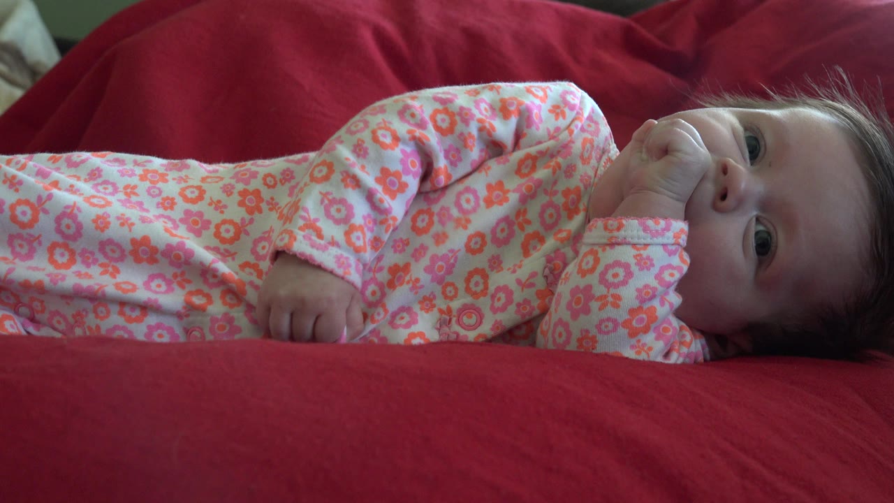 Baby lying on red bedding with a hand near the mouth.