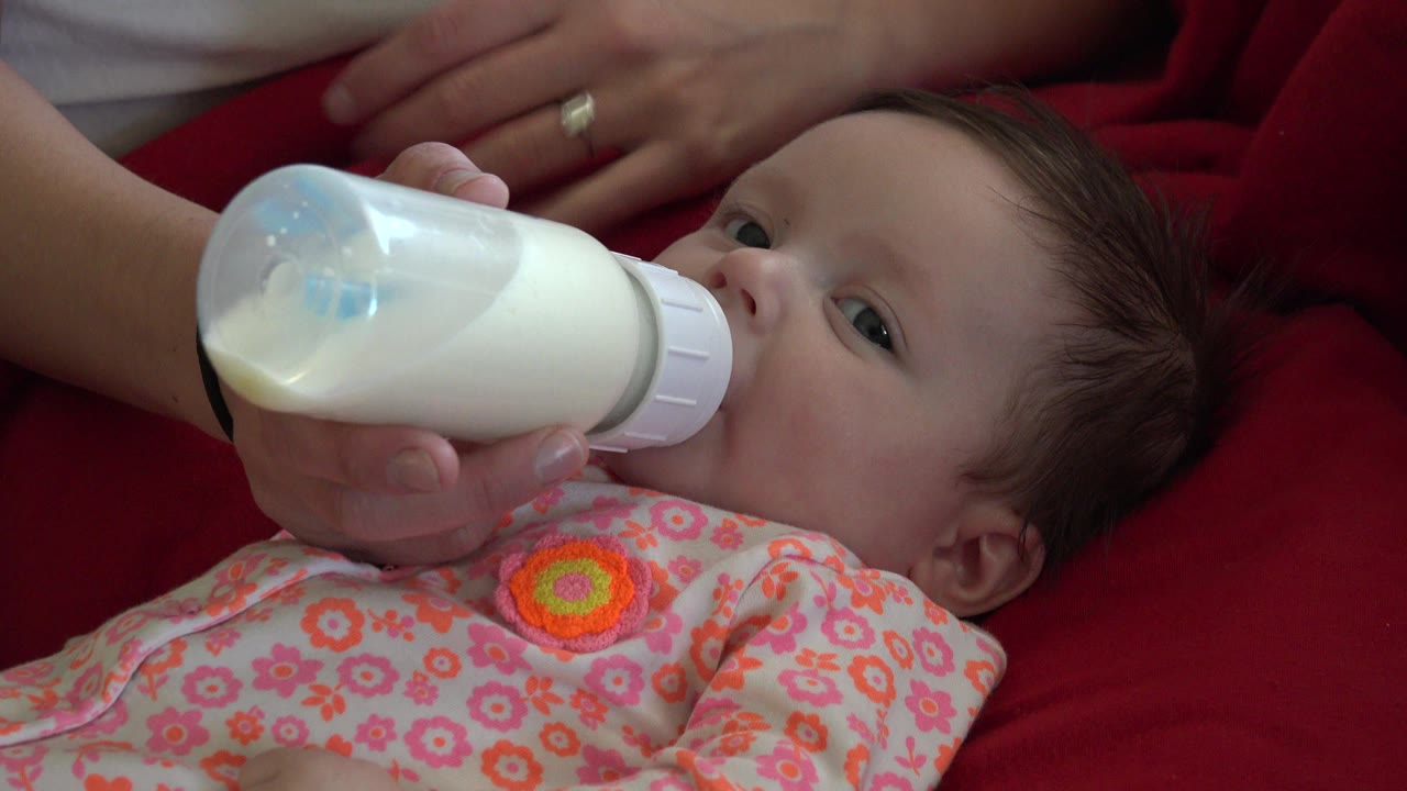 Baby lying on red fabric drinks from a milk bottle held by an adult hand.