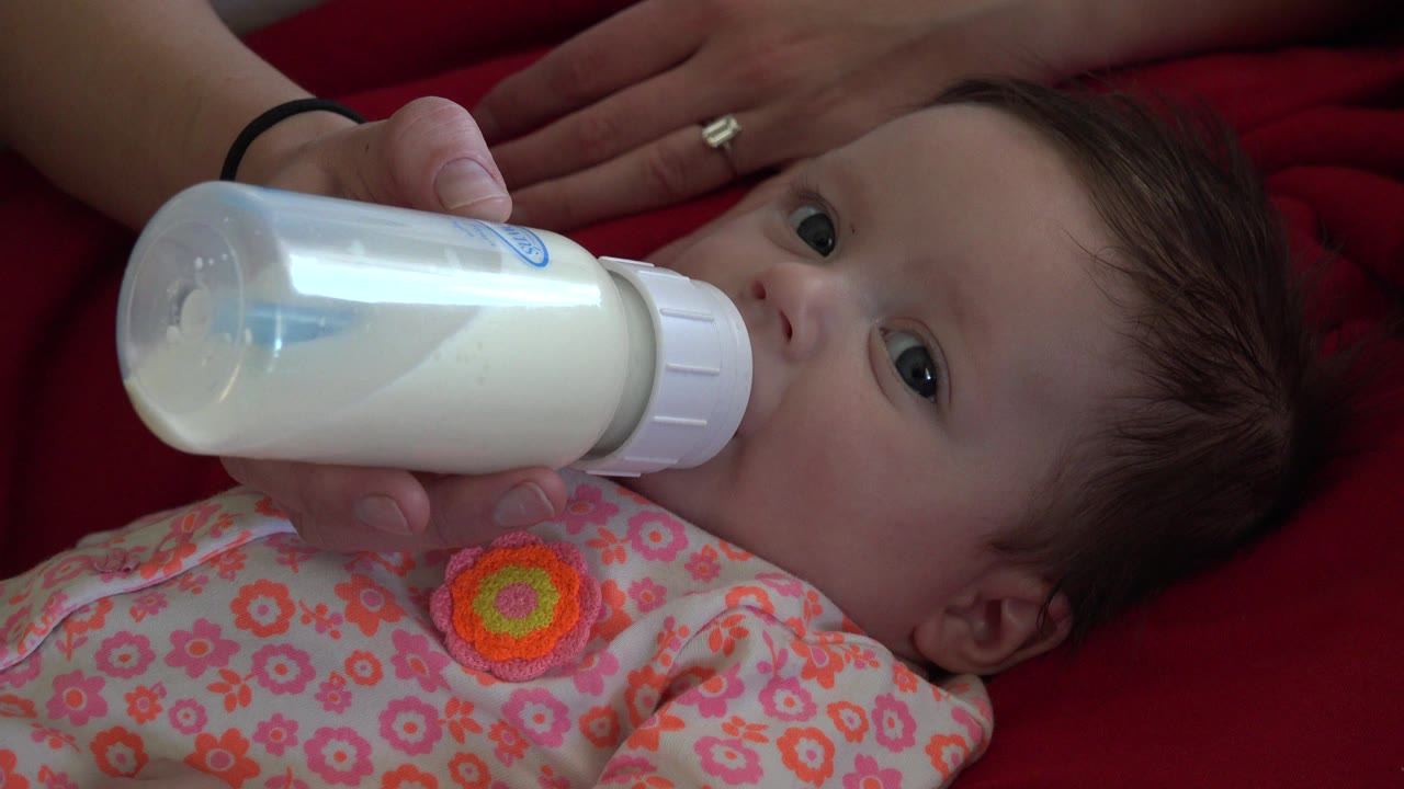 Baby drinking from a milk bottle held by an adult hand on red fabric.