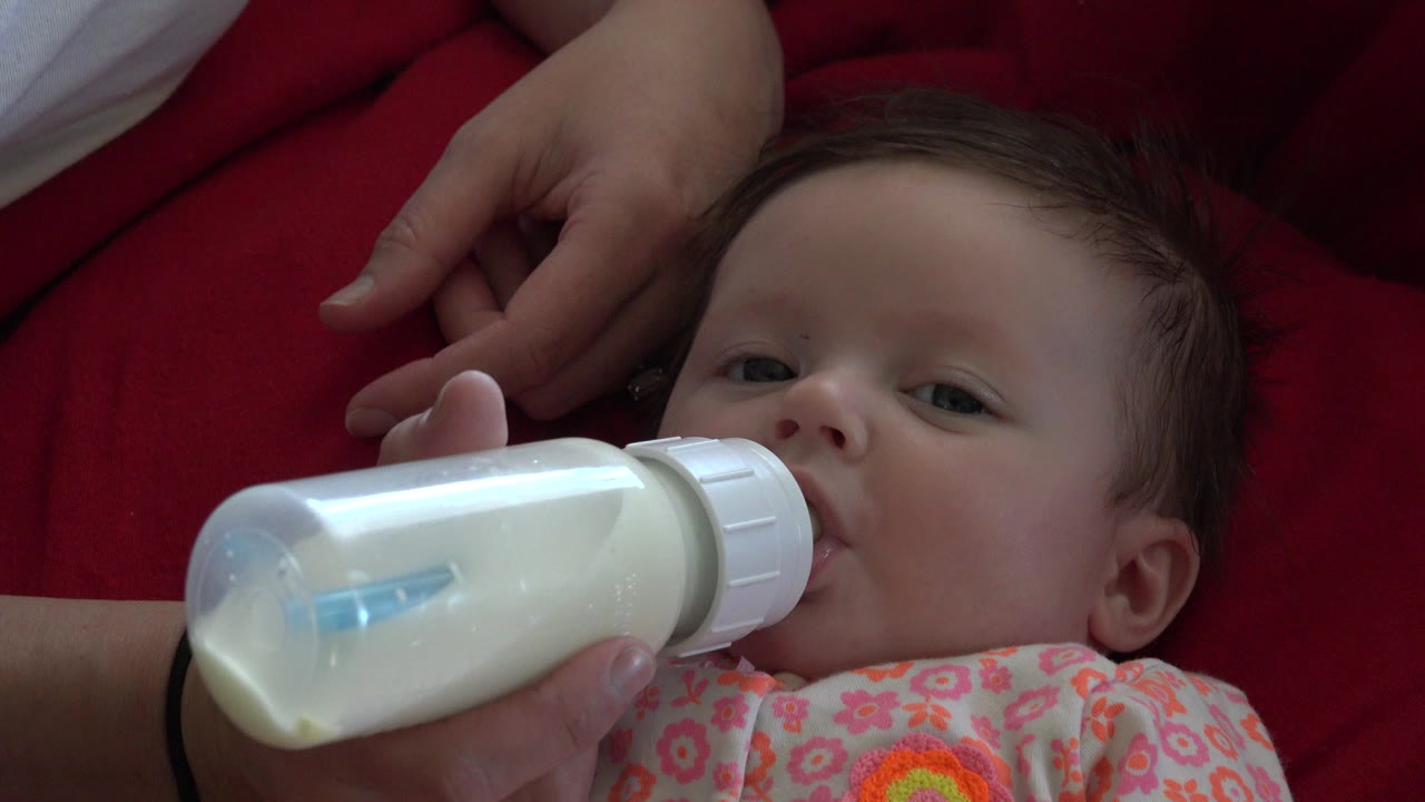 Baby lying on red quilt drinking from a milk bottle.