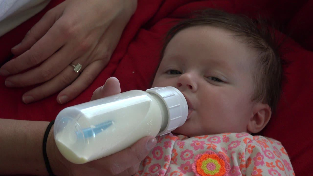 Baby being fed with a white bottle beside an adult hand on red fabric.