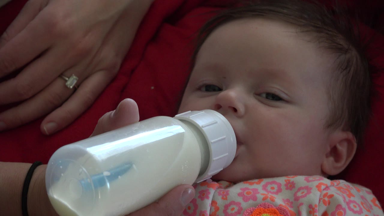At 0:00 in Westminster, Colorado, a baby drinks from a white bottle while an adult hand rests against red fabric behind the child.