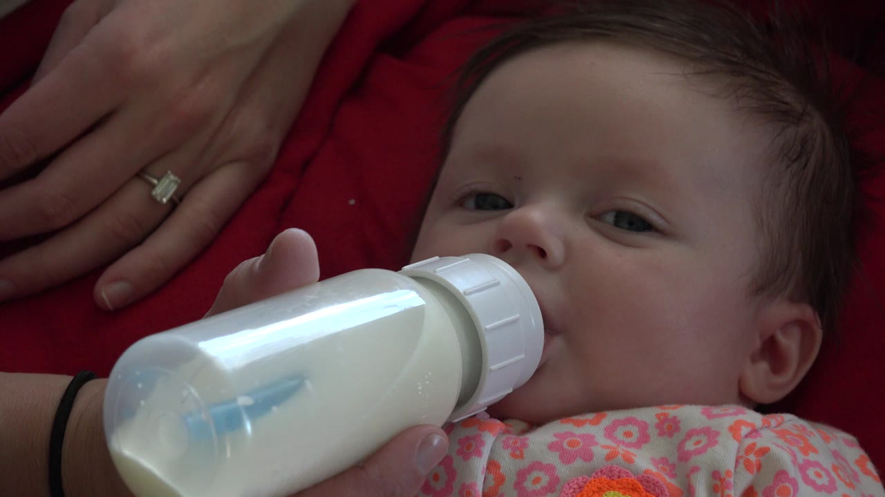 Baby lying against red fabric while drinking milk from a bottle.