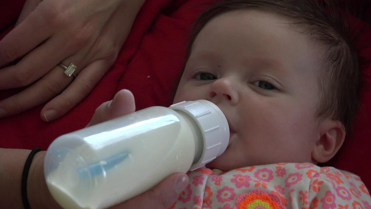 Infant drinking from a white bottle beside an adult hand on red fabric.