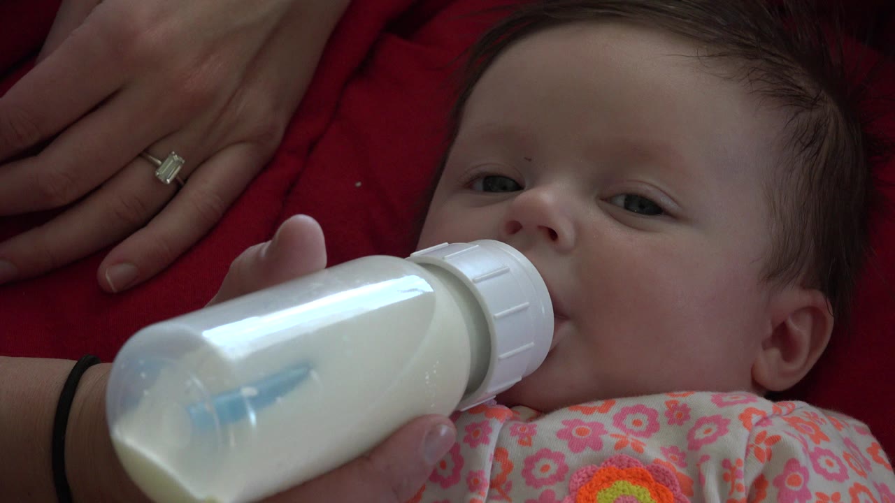 At 0:01 in Westminster, Colorado, the baby's eyes are open as the bottle covers the mouth and the adult hand remains above the head on red fabric.