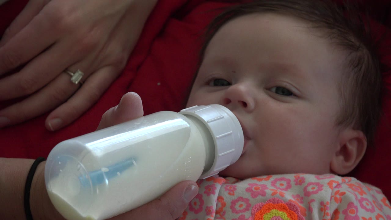Baby drinking from a milk bottle beside an adult hand on red fabric.