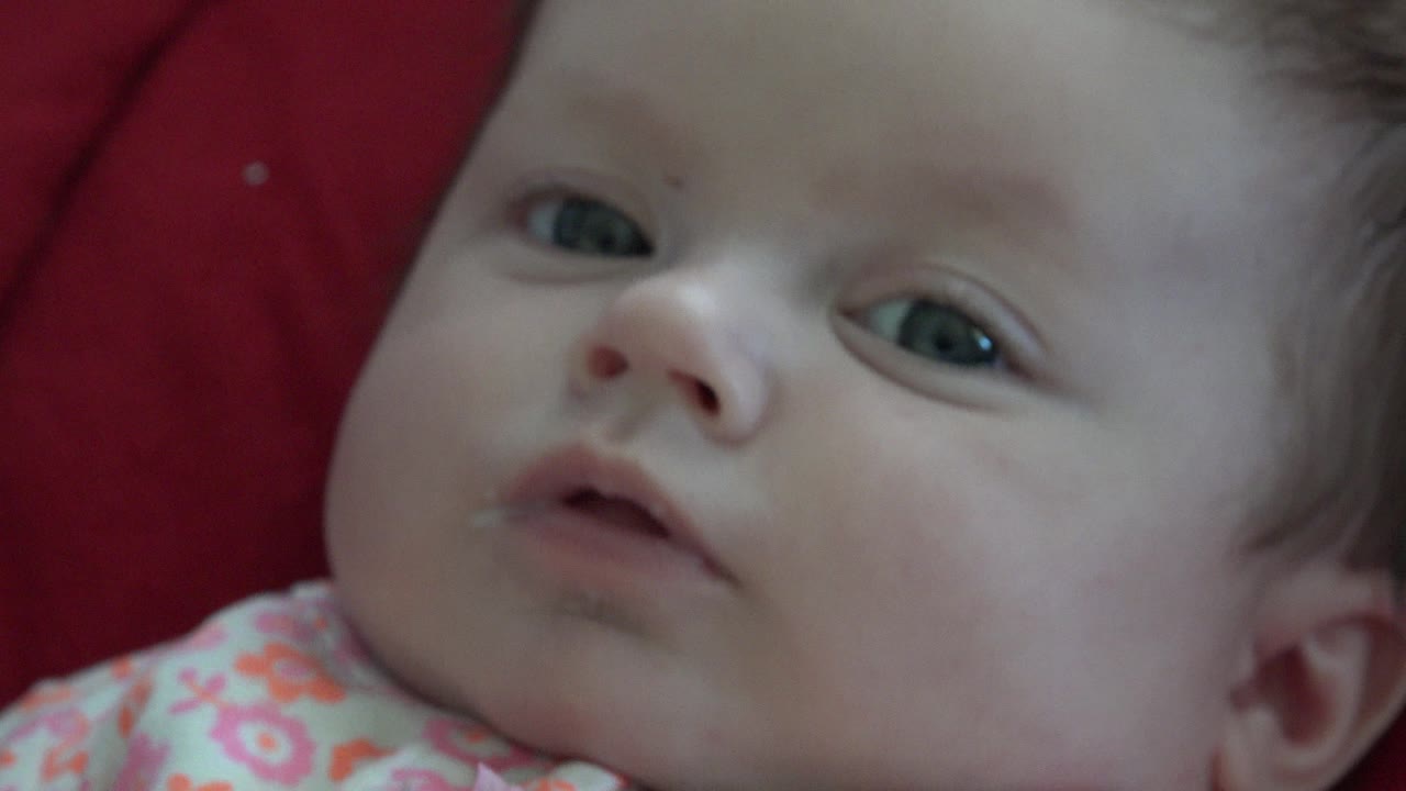 The baby remains in close-up on red fabric in Westminster, Colorado, with the mouth slightly open and the face filling most of the frame.