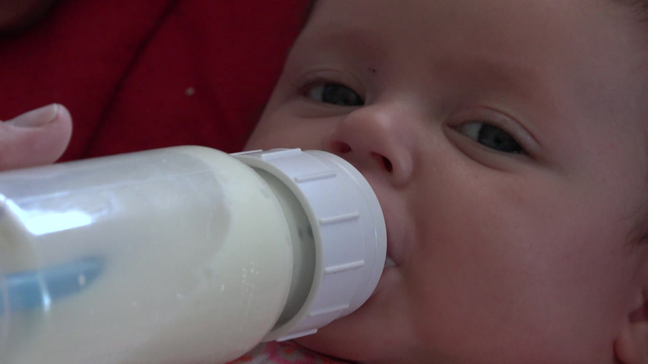 At 0:01, the bottle remains pressed to the baby's mouth while one eye looks toward the camera and the red cushion stays behind the face.