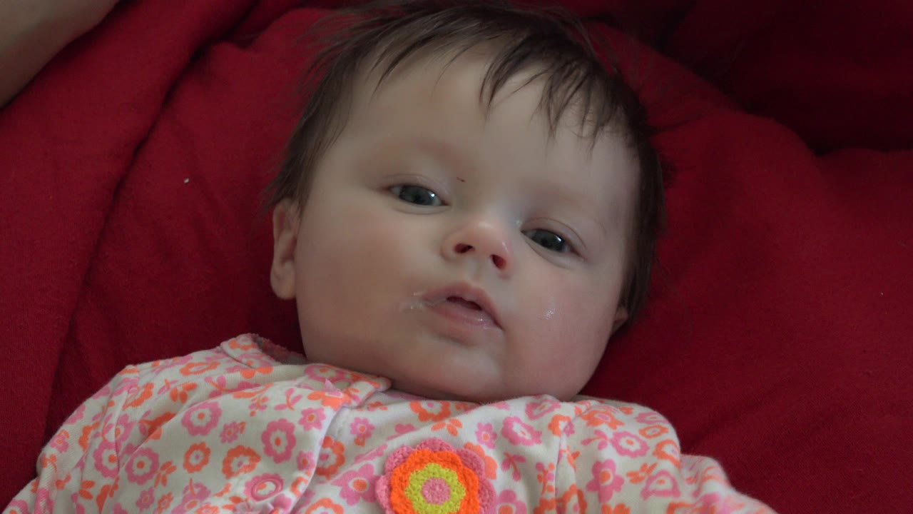 A baby lies on a red blanket in Westminster, Colorado, wearing a pink floral outfit and looking toward the close camera at the start of the clip.