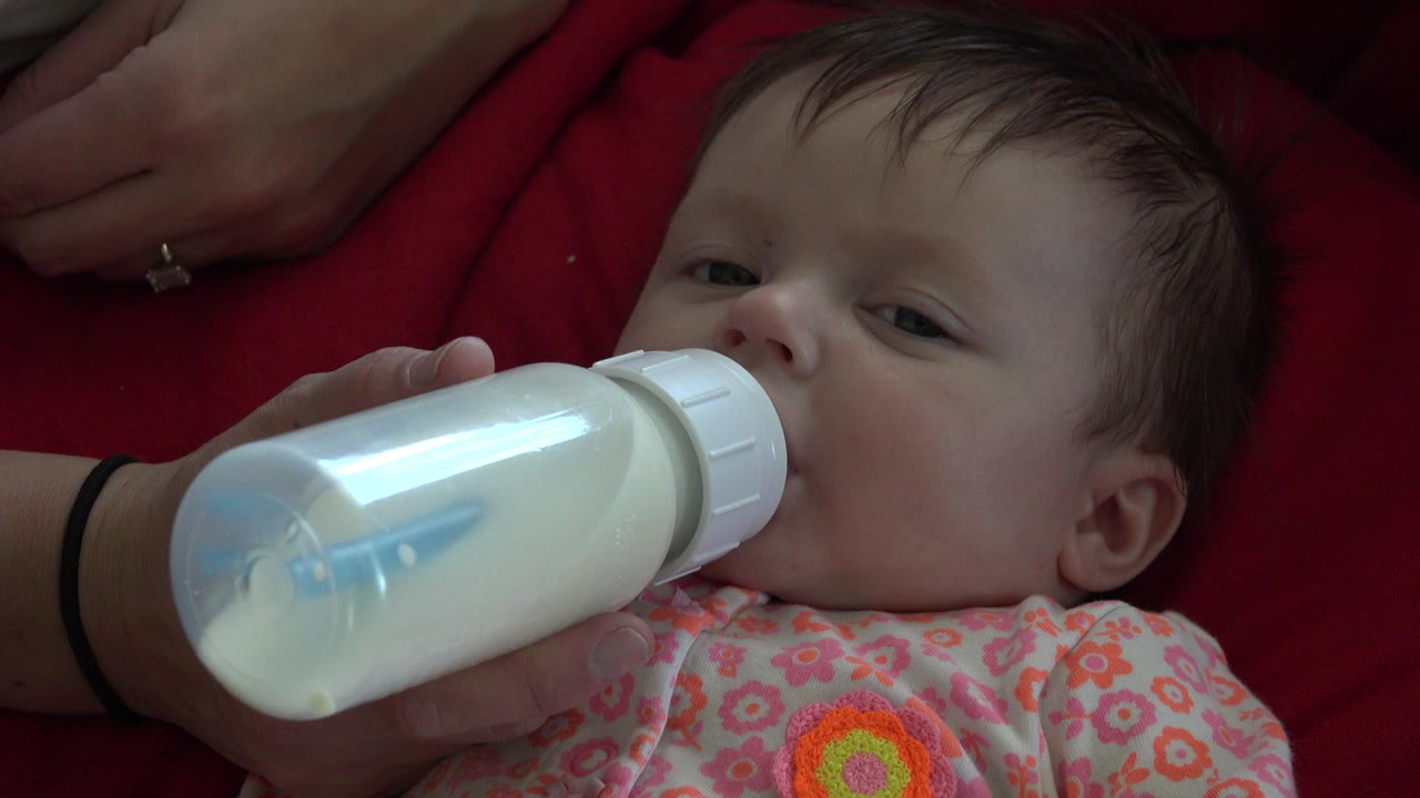 Baby lying on a red blanket drinking from a bottle held by an adult hand.