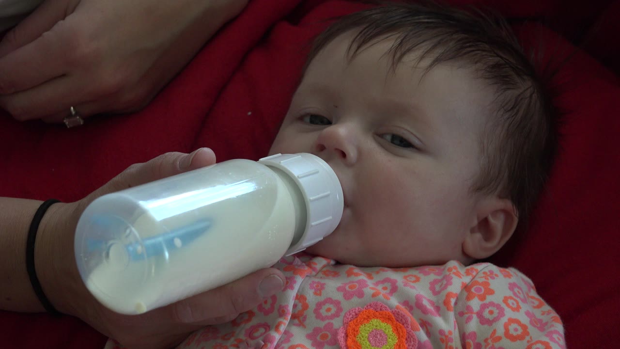 At 0:01 in Westminster, Colorado, the infant keeps the bottle at the mouth while the adult hand supports it from the lower left.