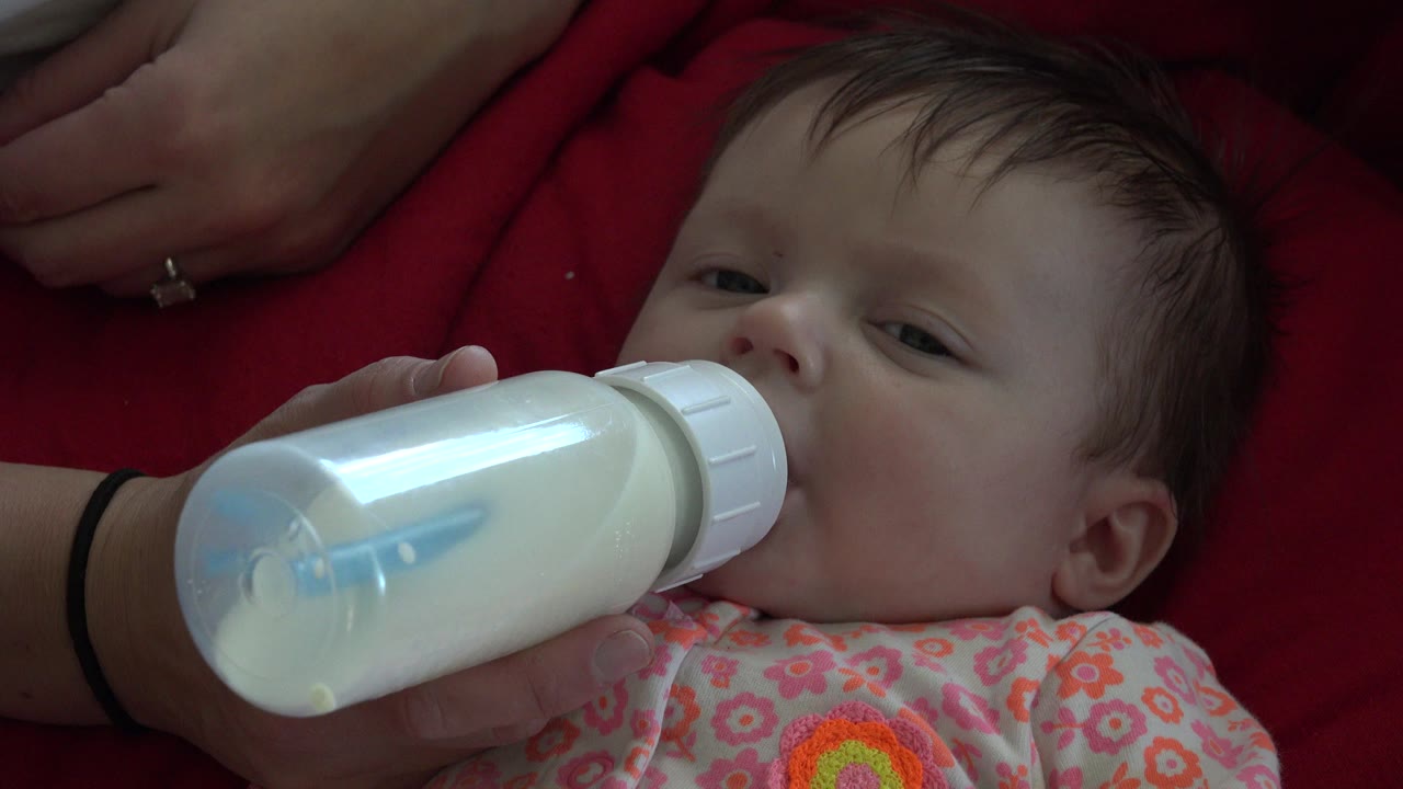 At 0:01, the bottle remains pressed to the infant's mouth as the baby's eyes look partly toward the camera against the red blanket.