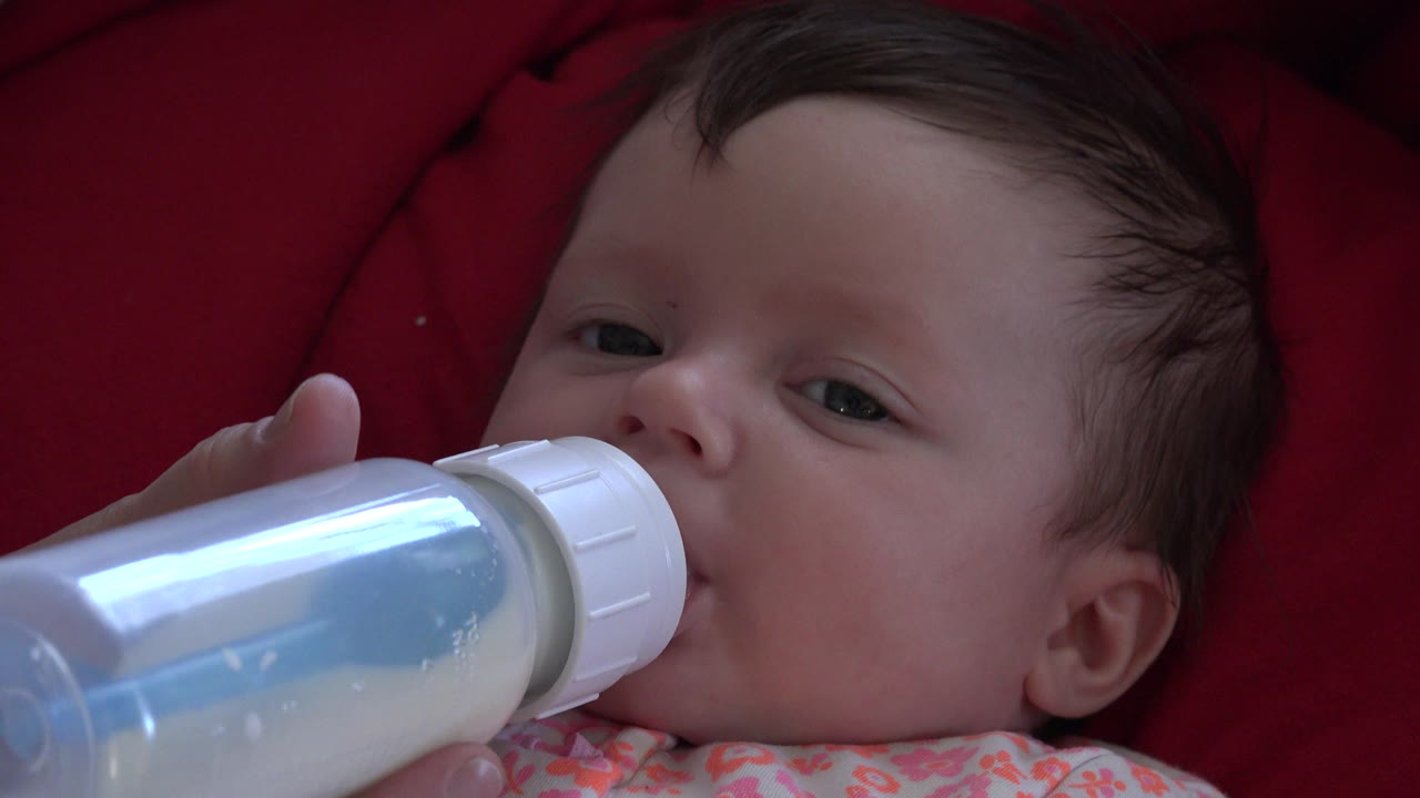 In Westminster, Colorado, the infant remains reclined on red fabric, eyes partly open, with the bottle still pressed to the mouth.