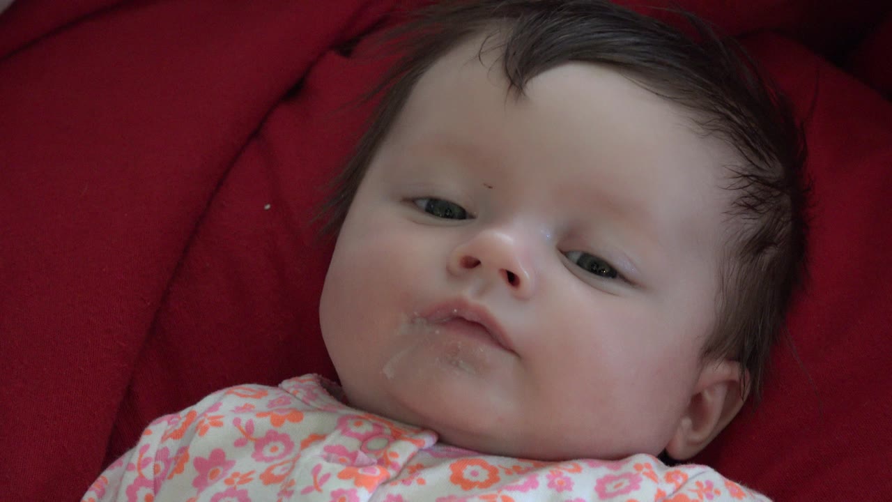 At 0:00, a baby lies on a red comforter in Westminster, Colorado, looking upward with a neutral expression and a floral outfit visible at the bottom of frame.