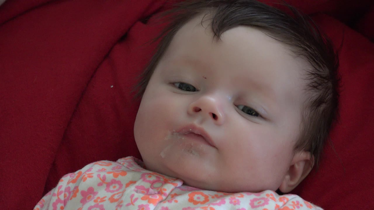 At 0:01, the baby's face remains centered against the red comforter, with dark hair, half-open eyes, and moisture visible around the lips.