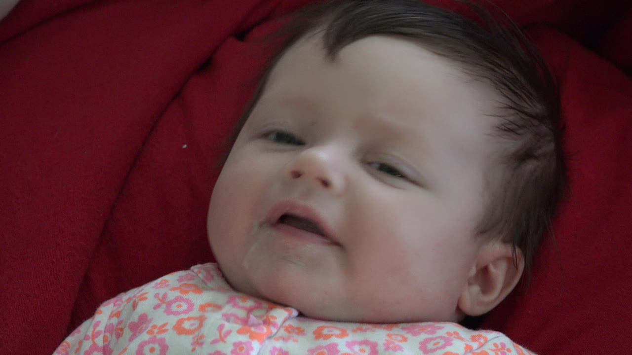 Baby in Westminster, Colorado remains framed close on the red cushion, with the mouth slightly open and the floral clothing visible along the lower edge.