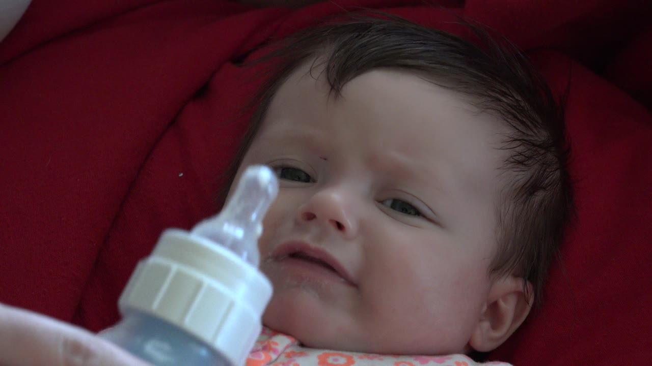 At 0:00, a baby in Westminster, Colorado lies on red cushioning as a translucent baby bottle is held near the face from the lower left.