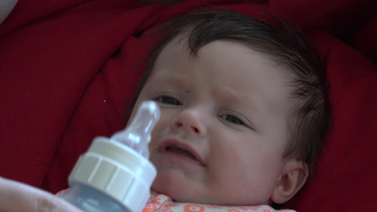 At 0:01, the baby's face remains centered on red cushioning while the bottle nipple is close to the mouth but not yet fully in place.