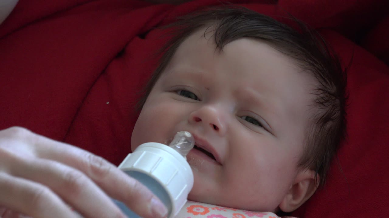 Adult hand holds a bottle to a baby’s mouth on red cushioning.