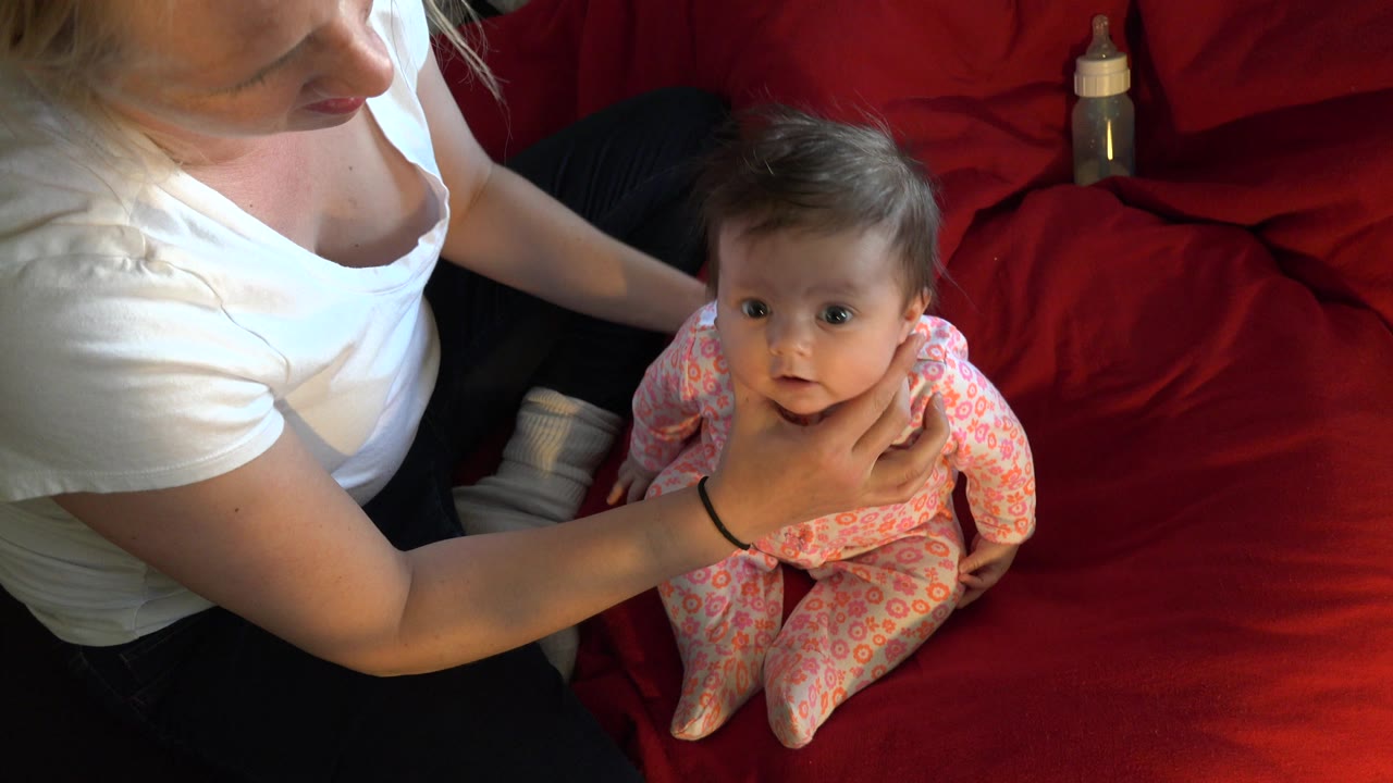 At 0:01 in Westminster, Colorado, the baby looks toward the camera as the woman’s hands hold the child upright on the red couch.