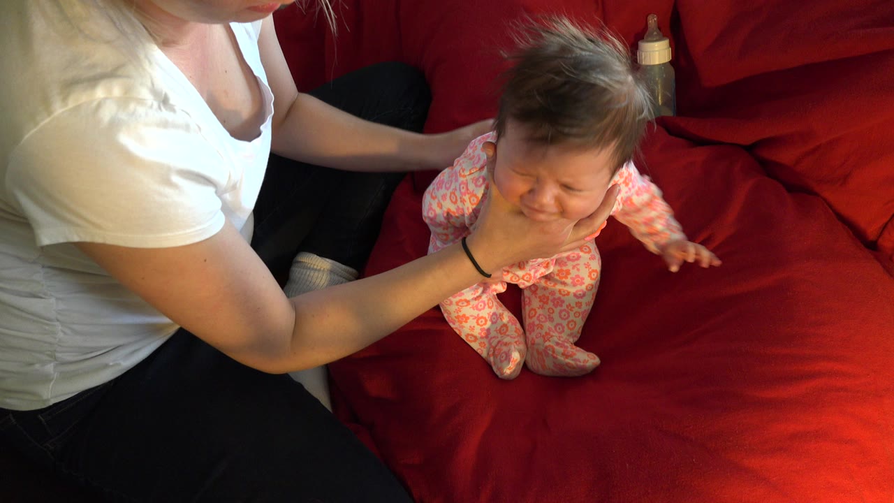 In Westminster, Colorado, an adult in a white shirt supports a baby in a pink sleeper standing or sitting on a red bed blanket at the start of the clip.