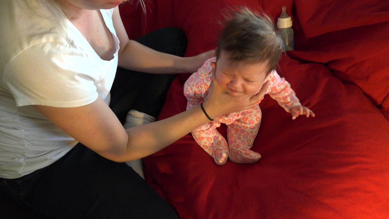 The adult remains seated at the left while holding the baby upright on the red blanket, with the infant's face turned toward the camera in the Westminster home interior.