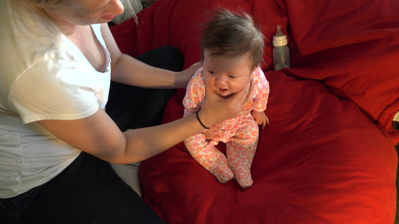 Woman supports a baby sitting on a red sofa.