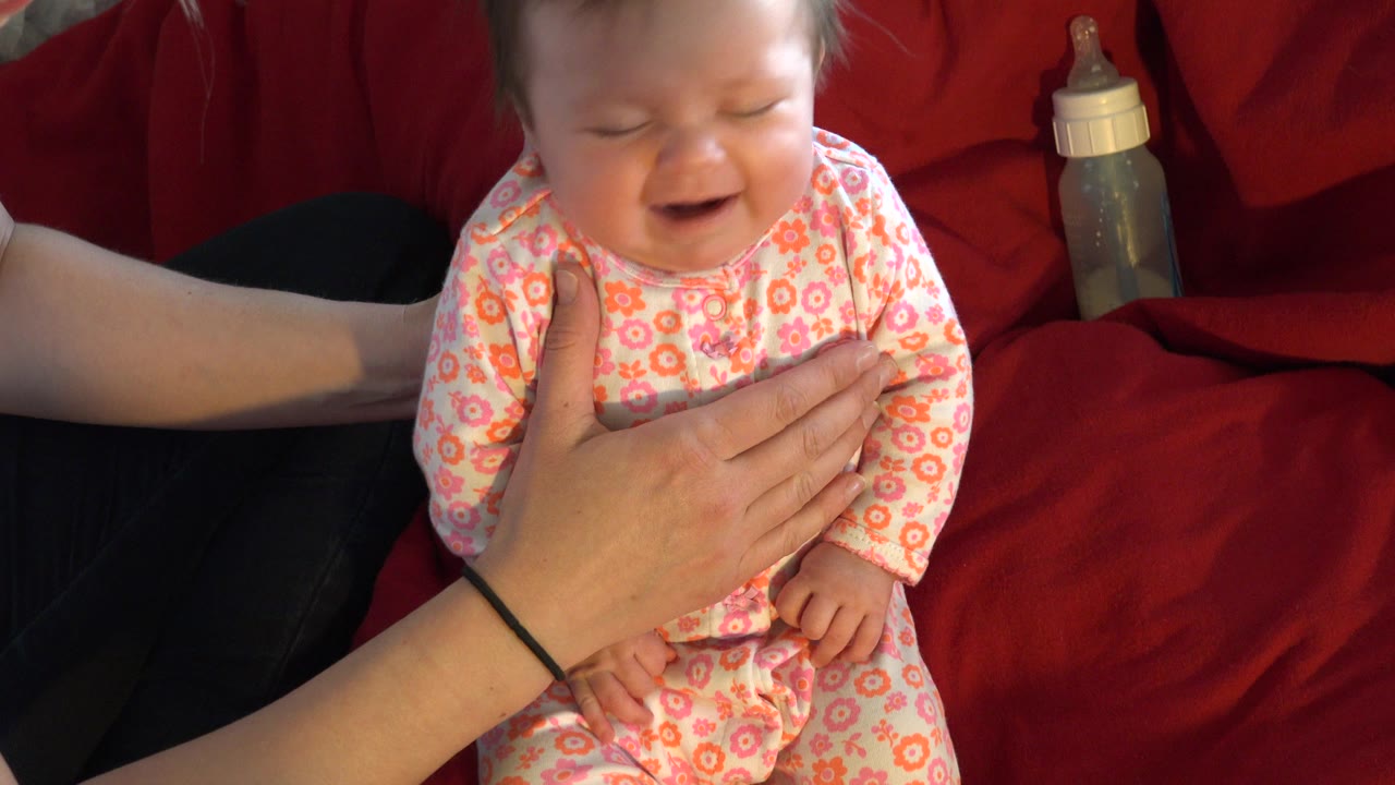 A baby in Westminster, Colorado sits on a red couch in a pink floral onesie while an adult hand steadies the infant's chest and a bottle rests behind them.