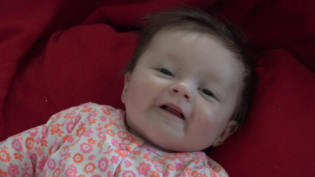 A baby in a floral outfit lies on a red blanket in Westminster, Colorado, facing the camera with a slight open-mouth expression at the start of the clip.