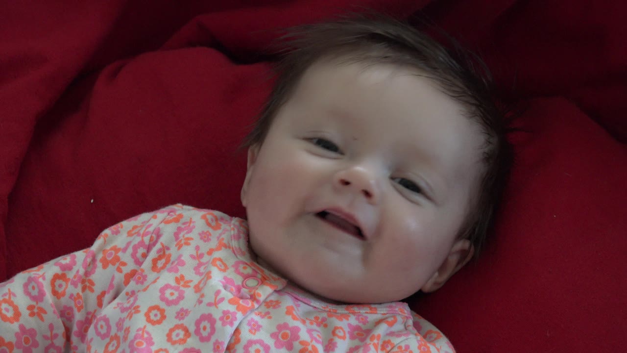 The baby remains on the red blanket in Westminster, Colorado, looking upward with eyes partly open and the floral outfit visible across the lower frame.
