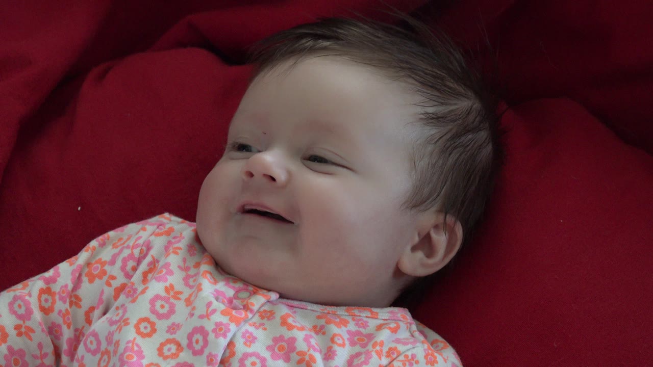 A baby in Westminster, Colorado lies on a red blanket at the start of the clip, wearing a pink floral outfit and looking upward with a small smile.
