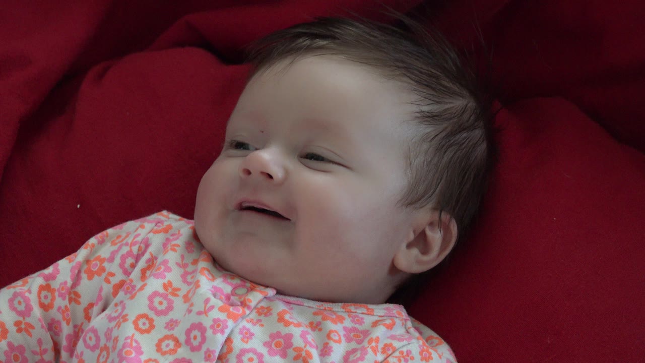 The baby remains on the red blanket in Westminster, Colorado, with the head turned slightly and the mouth opening into a smile.